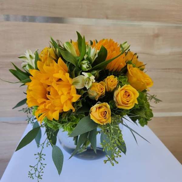 Low arrangement of yellow roses and gerbera daisies in a clear glass vase with green accents