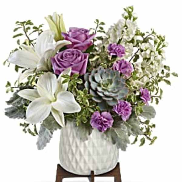 Arrangement of white lilies, lavender roses, purple carnations and a succulent in a white pot on wooden stand