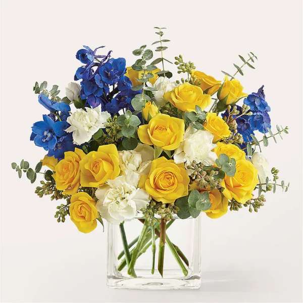 Yellow roses, blue flowers, and white carnations in a clear glass vase