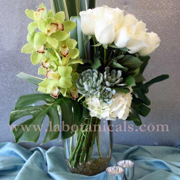 White roses and green orchids in a glass vase with tropical leaves