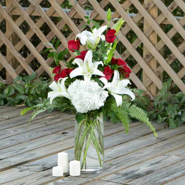 White lilies and red roses in a clear glass vase with white candles nearby.