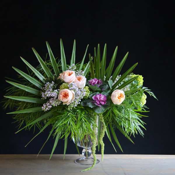 Peach roses and purple flowers in a glass vase with palm fronds