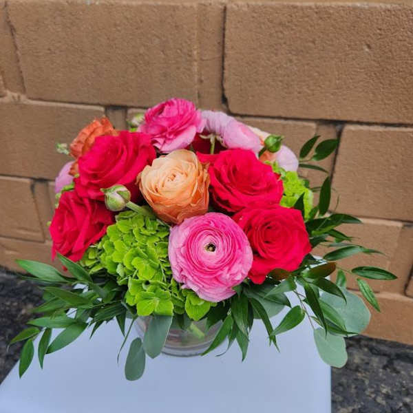 Short glass vase of pink roses, ranunculus, and green hydrangeas.