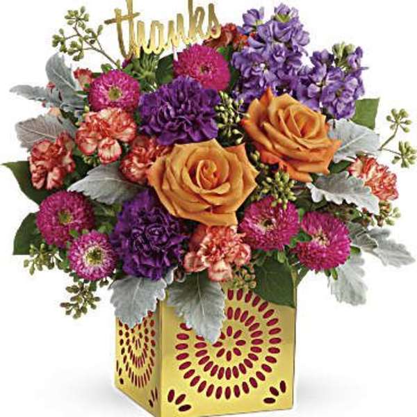 Bright mixed arrangement of orange roses, purple and pink flowers in a yellow cube vase with a gold “thanks” sign