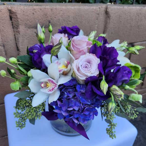 Low glass vase of purple and blue hydrangea, white orchids, and light pink roses in a compact arrangement