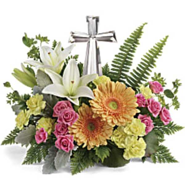 Low arrangement of white lilies, peach gerbera daisies, pink roses and yellow carnations with a silver cross.