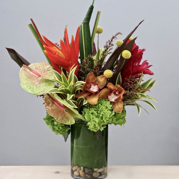 Tall tropical arrangement with red and orange blooms, orchids, and green hydrangea in a glass vase.