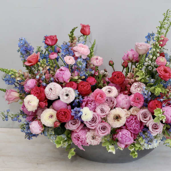 Low oval arrangement of pink and red roses, ranunculus, and peonies with blue delphinium in a gray bowl.