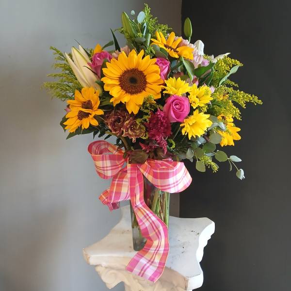 Mixed bouquet of sunflowers, roses, and lilies in a glass vase with a pink plaid ribbon.