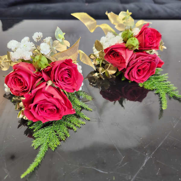 Pair of red rose and white accent flower wrist corsages with gold ribbon and fern accents on a reflective black surface.