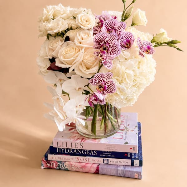 White roses, hydrangeas, and orchids arranged in a glass vase on stacked books