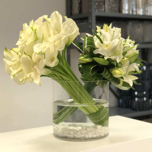 Modern arrangement of white calla lilies and lilies in a low glass vase with white stones