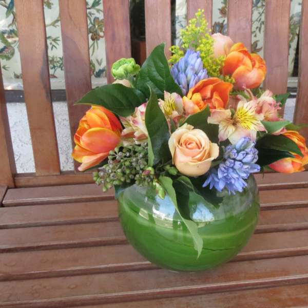 Mixed bouquet of orange, peach, and blue flowers in a green glass vase