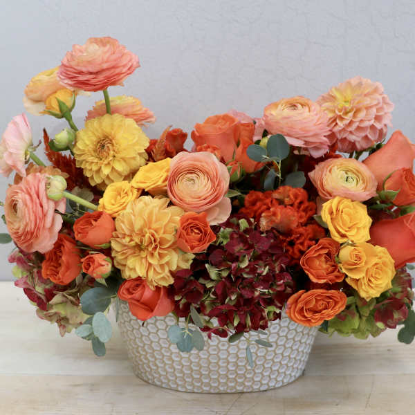 Low arrangement of orange, yellow, and peach flowers in a white patterned ceramic container