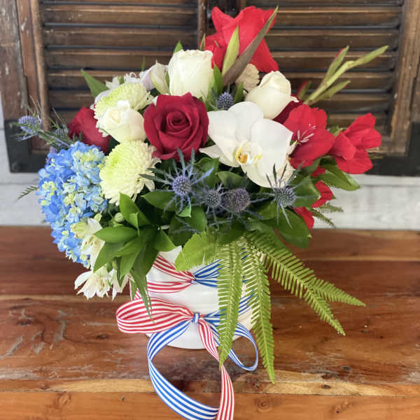 Red, white, and blue floral arrangement in a white vase with ribbon