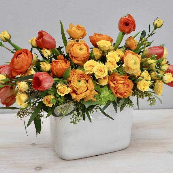 Low arrangement of orange ranunculus, yellow spray roses, and coral tulips in a white rectangular vase