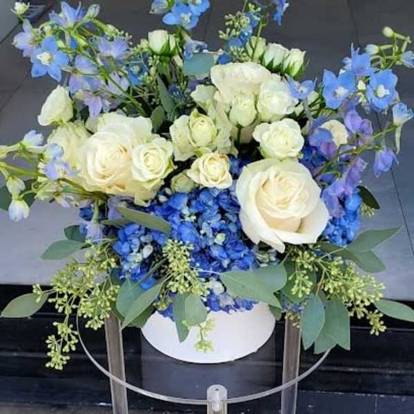 White roses and blue flowers arranged in a round white box on a clear stand