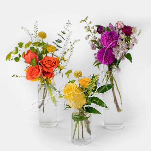 Trio of small mixed flower arrangements with yellow, orange, and purple blooms in clear glass bud vases
