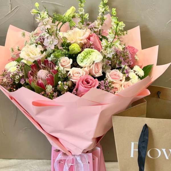 Large pink wrapped bouquet with pastel roses, protea, and mixed flowers beside a kraft shopping bag