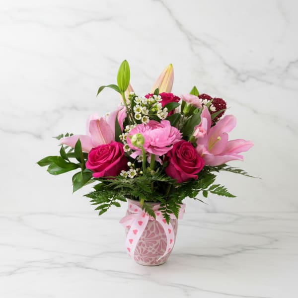 Pink roses, lilies, and daisy-style blooms in a pink patterned vase with a heart ribbon