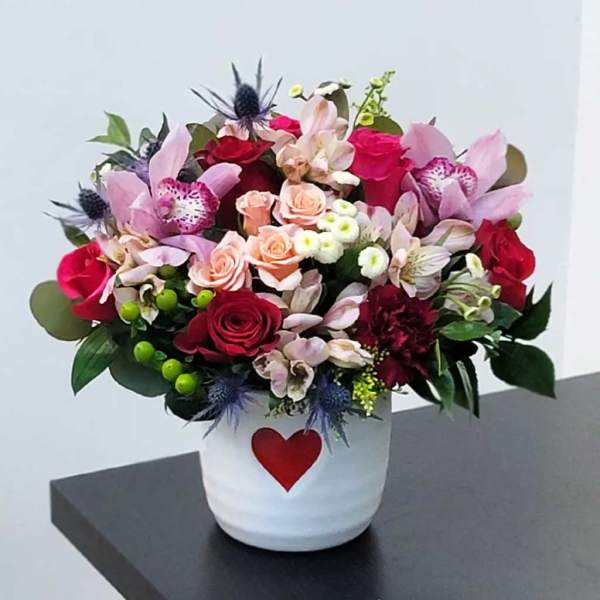 Mixed pink and red flower arrangement in a white pot with a red heart
