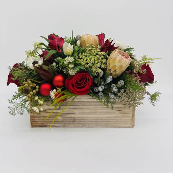 Low red and white flower arrangement with protea and roses in a wooden rectangular box with red ornaments.
