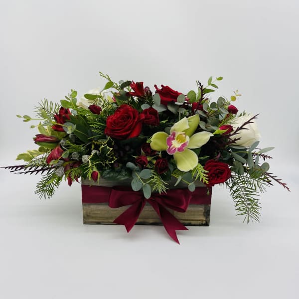 Low rectangular arrangement of red roses, white blooms, and green orchids in a wooden box with a red bow