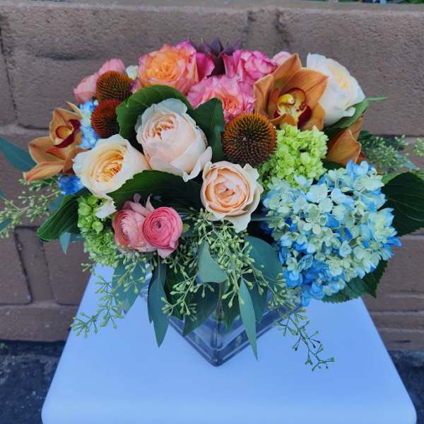 Low mixed arrangement of peach roses, blue hydrangeas and orchids in a square glass vase