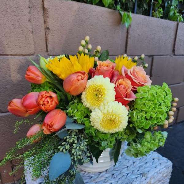 Low arrangement of orange tulips, yellow sunflowers, roses, mums and green hydrangea in a white vase