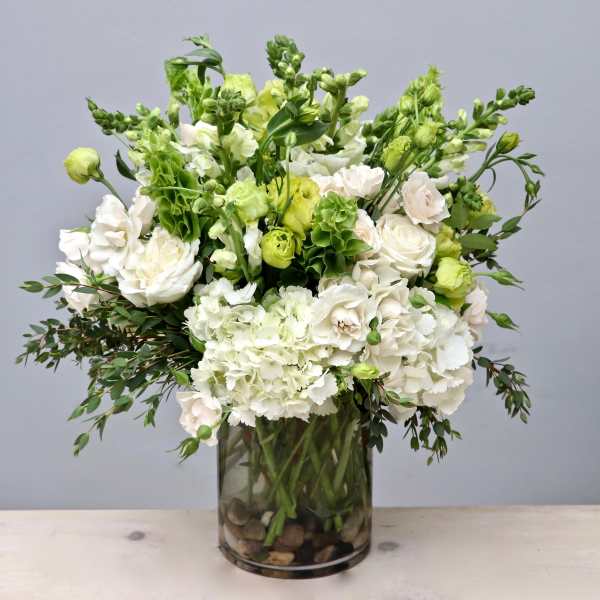 Tall arrangement of white and green flowers in a clear glass cylinder vase with stones at the base