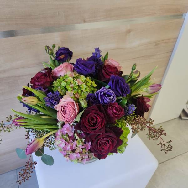 Low round arrangement of red roses, purple lisianthus, tulips and hydrangeas in a clear glass vase