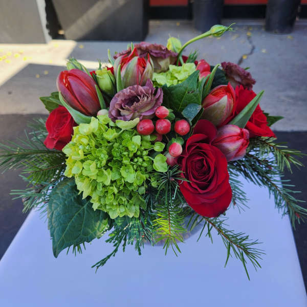 Short arrangement of red roses, tulips, green hydrangea, berries and lisianthus in a glass vase with evergreen sprigs.