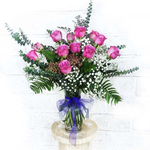 Pink roses in a glass vase with white filler flowers and purple ribbon