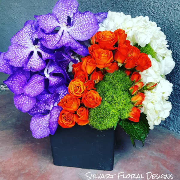 Purple orchids, orange roses, and white hydrangeas in a black square vase