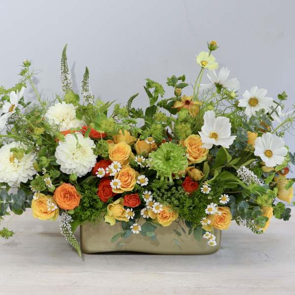 Low arrangement of yellow and orange roses with white blooms in a rectangular gold vase