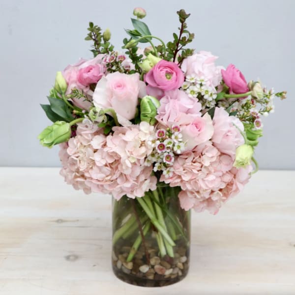 Pastel pink hydrangeas, roses, and ranunculus arranged in a clear glass vase with pebbles at the base