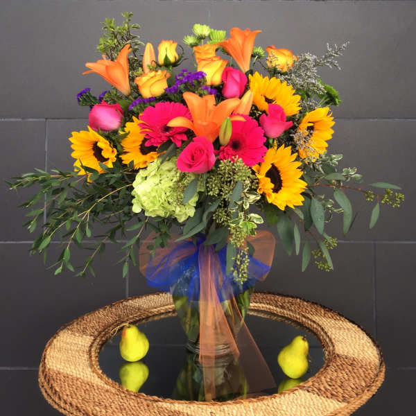 Bright mixed bouquet with sunflowers, roses, lilies, and gerbera daisies in a glass vase.