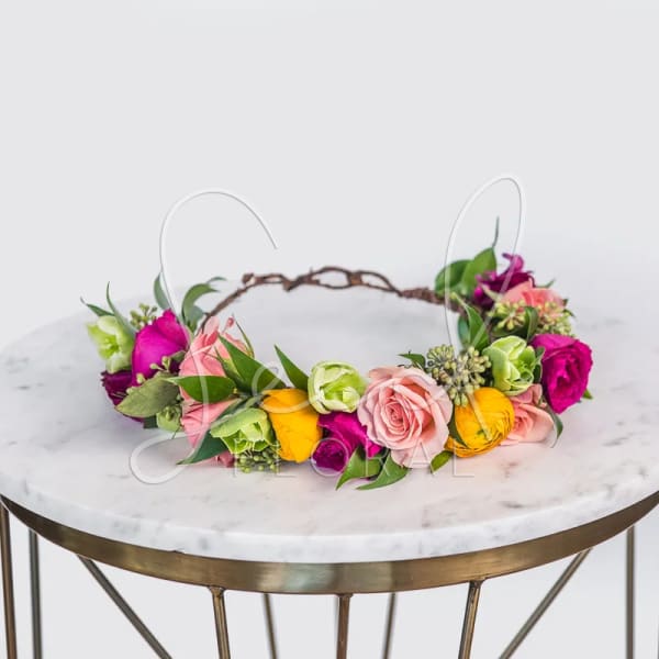 Colorful flower crown with pink roses, yellow blooms and green buds on a vine base, displayed on a round marble table.