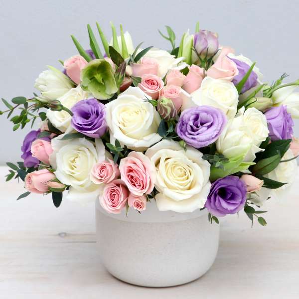 Round arrangement of white roses, pink roses, and lavender lisianthus in a white ceramic pot