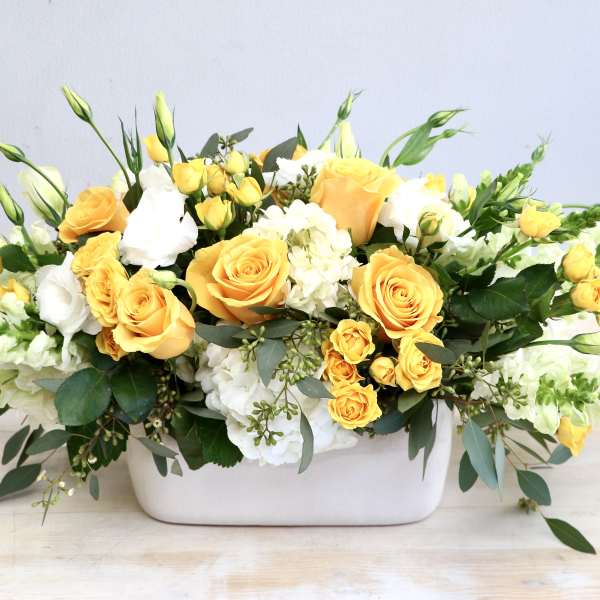 Low arrangement of yellow roses and white flowers in a rectangular white container
