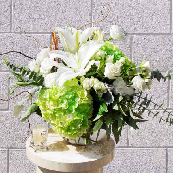 White lilies and green hydrangeas in a glass vase with a candle beside it