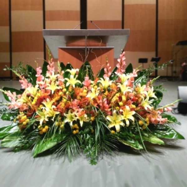 Low, wide arrangement of yellow lilies and peach flowers with large green foliage in front of a stage podium