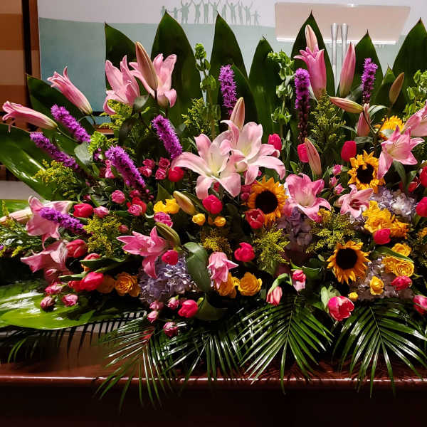 Large horizontal event arrangement of pink lilies, tulips, sunflowers, and mixed blooms with bold tropical foliage