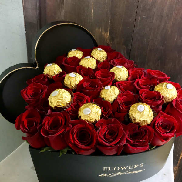 Red roses arranged with gold-wrapped chocolates in a black heart-shaped box