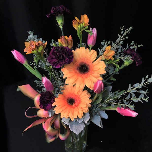 Orange gerbera daisies with pink tulips and dark purple carnations in a glass vase