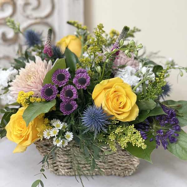 Mixed flower arrangement in a woven basket with yellow roses and purple blooms