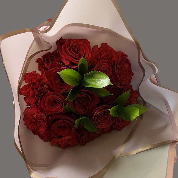 Hand-tied bouquet of red roses and red carnations with bright green leaves wrapped in white paper.