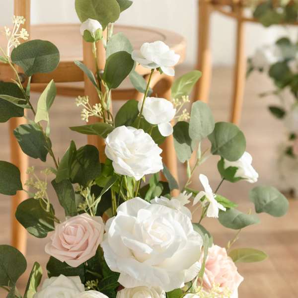 Floor arrangement of white and blush roses with abundant greenery beside wooden chairs
