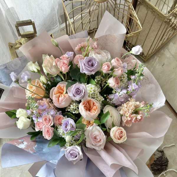 Pastel bouquet of pink, peach, and lavender roses with white accents wrapped in soft mauve paper