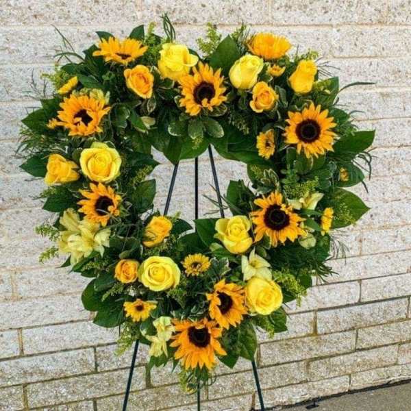 Heart-shaped wreath of yellow roses and sunflowers on a stand against a brick wall.